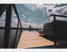 Eine offene Holzplattform auf einem See, umgeben von einem Geländer, mit einem Sofasitzbereich auf der rechten Seite und einer DJ-Station in der Mitte. Der Himmel ist leicht bewölkt, und im Hintergrund erstreckt sich eine bewaldete Uferlinie.