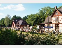 Bahnhofsgebäude im Fachwerkstil mit dunklem Dach inmitten einer grünen, von Bäumen umgebenen Landschaft; davor Sitzgelegenheiten mit Tischen, an denen sich viele Menschen versammelt haben. Ein Schild mit der Aufschrift „Kaiserbahnhof“ ist im Vordergrund sichtbar.