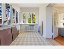 Heller, leerer Innenraum mit großen Fenstern auf zwei Seiten, einer niedrigen Fensterbank und Blick ins Grüne. In der Mitte steht ein Heizkörper unter dem Fenster, der Boden ist mit hellgrau-weißem Rautenmuster gefliest, und rechts führt eine offene Tür in einen angrenzenden Raum mit Holzboden.