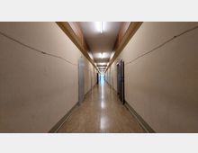 Langer, schmaler Flur mit cremefarbenen Wänden und linoleumartigem Bodenbelag, beleuchtet durch an der Decke montierte Neonröhren. Auf beiden Seiten befinden sich geschlossene graue Türen, und am Ende des Flurs ist eine verglaste Tür zu sehen.