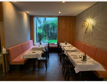 Ein modernes Restaurant mit dezenter Beleuchtung, Holzwänden und grauen Betonakzenten. Es gibt zwei Reihen von Tischen mit weißen Tischdecken, schwarzen Stühlen und einer langen, orangefarbenen Polsterbank; im Hintergrund ist ein großes Fenster mit Blick auf einen begrünten Außenbereich.