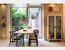 Heller Essbereich mit einem großen Holztisch, umgeben von schwarzen Stühlen mit geflochtenen Sitzflächen. Im Hintergrund eine Fensterfront mit Blick ins Freie, flankiert von Backsteinwänden, einem hohen Vitrinenschrank und Wanddekorationen.