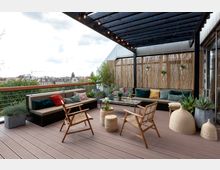 Moderne Dachterrasse mit überdachtem Sitzbereich, der durch Holzmöbel, eine lange gepolsterte Bank mit bunten Kissen und Dekoelemente wie Pflanzen und Korbmöbel gestaltet ist. Im Hintergrund bietet ein Bambus-Sichtschutz Privatsphäre, während die umliegenden Dächer der Stadt sichtbar sind.