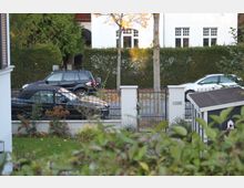 Gepflegter Vorgarten mit Hecken, Rasen und Sträuchern, im Vordergrund ein Zaun mit einem Tor aus Metall. Dahinter stehen parkende Autos auf einer ruhigen Straße vor einem weißen Wohngebäude.