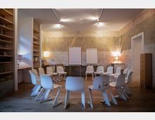 Ein heller Seminarraum mit abgenutzten Wänden, Holzfußboden und kreisförmig angeordneten weißen Stühlen. Im Hintergrund stehen zwei Flipcharts vor einer Wand, seitlich befinden sich Holzregale, Lampen und ein geschlossener Raumzugang.