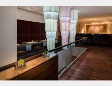 Ein modernes Lounge-Interieur mit hängenden, mehrteiligen Lampen aus hellem Stoff. Rechts ist eine Bar mit dunklen Stühlen vor einer beleuchteten Wand, während links Sitzbereiche mit braunen Polsterwänden und kleinen, dekorativen Elementen auf den Tischen zu sehen sind.