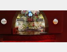 Eine Bar mit gebogenem Torbogen und einer geschmückten Rückwand mit tropischem Pflanzenmuster und Tierillustrationen. Auf den Regalen stehen Flaschen und Gläser, während die Beleuchtung eine warme, rote Atmosphäre erzeugt.