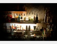 Eine Bar mit einer Theke, auf der Gläser, Flaschen und Barkeeper-Utensilien stehen. Im Hintergrund sind Regale mit aufgereihten Spirituosenflaschen, beleuchtet von einer warmen Lichtquelle, und eine rau anmutende Wand zu sehen.