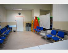 Ein einfacher Wartebereich mit blauen Stühlen, einer geschlossenen weißen Tür, einer Uhr an der Wand und diversen gelagerten Gegenständen, darunter rote und gelbe Rettungsgeräte, sowie ein provisorisches weißes Tuch an der rechten Seite. Der Raum hat helle Wände und einen beigen Boden.