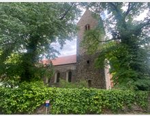 Historische Kirche aus Feldstein mit Turm und roten Ziegeldach, umgeben von hohen Bäumen und einer mit Efeu bewachsenen Steinmauer. Der Himmel ist leicht bewölkt, und ein Verkehrsschild steht vor der Mauer.