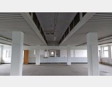 Leerer, rechteckiger Raum mit weiß gestrichenen Wänden, mehreren tragenden Säulen und einem abgehängten Deckenbereich mit eingebauten Oberlichtern. Die Wände sind teilweise mit weißen Fliesen verkleidet, und der Raum wirkt unbeheizt mit Spuren von Verfall an der Decke.