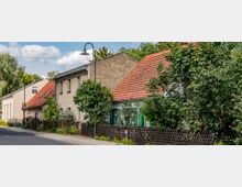 Straßenszene in einer ländlichen Umgebung mit historischen Wohnhäusern, teils aus Backstein, und roten Ziegeldächern. Vor den Häusern stehen Bäume, Büsche und Holzzäune, daneben führt eine asphaltierte Straße entlang.