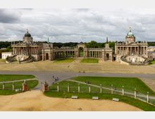 Historische barocke Gebäudeanlage mit zwei symmetrischen Bauwerken, verziert mit Kuppeln, Säulen sowie geschwungenen Treppen, verbunden durch eine halbkreisförmige Kolonnade. Vordergrund zeigt eine weitläufige Wiese, Gehwege, ein schmiedeeisernes Tor und vereinzelt spazierende oder radfahrende Personen.