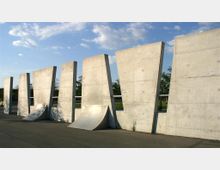 Eine Reihe großer, schräg stehender Betonwände mit integrierten Skate-Rampen davor, in einer offenen, parkähnlichen Umgebung bei klarem Himmel. Im Hintergrund sind Bäume und ein weitläufiger Rasen zu sehen.