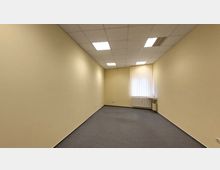 Leerer Raum mit beigen Wänden, grauem Teppichboden und einer Rasterdecke mit eingebauten Leuchtstofflampen. Am Ende des Raums befindet sich ein Fenster mit vertikalen Jalousien sowie ein Waschbecken mit einem kleinen Wandspiegel.