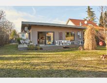 Einstöckiges, modernes Haus mit Flachdach, großer Terrasse und bodentiefen Fenstern. Im Vordergrund ein Garten mit Rasenfläche, dekorativen Pflanzen und einer Heuhaufen-Struktur.