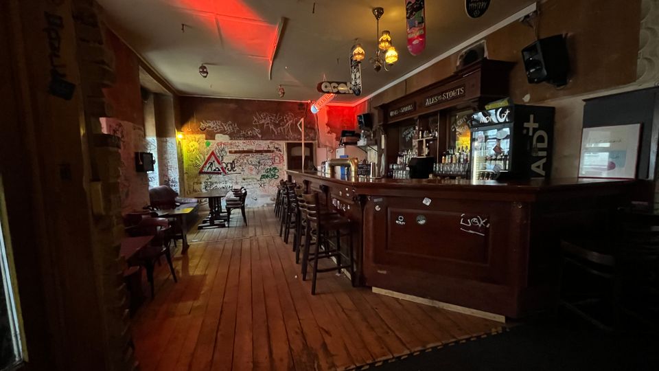 Innenansicht einer schmalen Bar mit langem Holztresen und Barhockern, Holzfußboden sowie kleinen Tischen an der linken Wand. Gedämpftes Licht mit roten Akzenten, Graffiti an den Wänden und Skateboards als Deckendekoration prägen den Raum; hinter dem Tresen stehen Regale mit Flaschen und ein beleuchteter Kühlschrank.