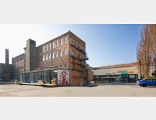 Backstein-Gebäudekomplex mit industriellem Stil, bestehend aus einem mehrstöckigen Bau mit Feuertreppe an der Fassade. Im Vordergrund befindet sich ein kleiner moderner Anbau mit Glasfront und einem gemalten Hahnmotiv; daneben ein flaches Gebäude mit farbigen Schriftzügen auf dem Dach.