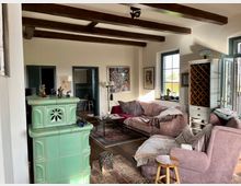 Gemütliches Wohnzimmer mit Holzbalkendecke, einem grünen Kachelofen im Vordergrund und einem Sitzbereich mit beigen Sofas und Sessel. Im Hintergrund sind offene Türen, große Fenster, ein Weinschrank und diverse Dekorationsobjekte sichtbar.