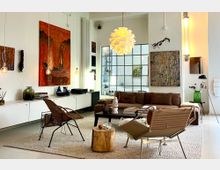 Modern living room with white walls, a large gridded window, and contemporary art pieces above a low white sideboard and brown sofa. A sculptural pendant light hangs over a rug with mixed lounge chairs and a coffee table, with minimalist floor lamps and decorative objects throughout.