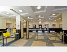 Eingangsbereich eines öffentlichen Gebäudes mit Sicherheitskontrolle, bestehend aus einem Metalldetektor, Glasdrehsperren und Geländern. Der Raum ist hell beleuchtet, mit einer abgehängten Decke, Fliesenboden und Hinweisschildern an den Wänden.