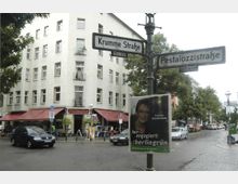 Straßenecke in einer städtischen Umgebung mit einem mehrstöckigen Wohngebäude und dem Restaurant "Thomas-Eck" mit roten Markisen im Erdgeschoss. Straßenschilder weisen auf die "Krumme Straße" und die "Pestalozzistraße" hin, Bäume säumen die Straßen, und im Vordergrund ist ein Wahlplakat angebracht.