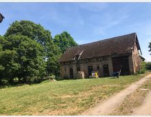 Ein altes Backsteingebäude mit einem verwitterten Ziegeldach steht auf einer Wiese, umgeben von großen, grünen Bäumen. Eine Schubkarre und landwirtschaftliche Geräte befinden sich vor dem Gebäude, rechts davon führt ein unbefestigter Weg entlang.