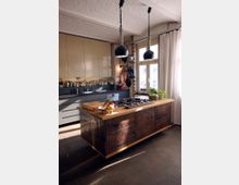 Moderner Küchenraum mit einer zentralen Kücheninsel aus dunklem Holz und einer hellen Holzarbeitsplatte. Im Hintergrund befinden sich helle, grifflose Schränke, ein Fenster mit weißen Vorhängen und offen sichtbare Backsteinwände, die industriellen Charme verleihen.