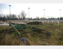 Eine Pferderennbahn mit weißen Zaunabgrenzungen und Flutlichtmasten im Hintergrund, ein Trainingswagen mit einem Pferd auf der Bahn. Im Vordergrund liegt eine Wiese mit gealterten landwirtschaftlichen Geräten und Metallrahmen.