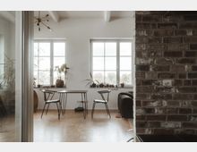 Minimalistisch eingerichtetes Wohnzimmer mit Holzboden, einem kleinen Tisch und zwei Stühlen vor großen Fenstern. Rechts im Bild ist eine gemauerte Ziegelwand zu sehen, links sind Zimmerpflanzen und ein Sofa im Hintergrund sichtbar.