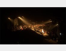 Eine dunkle Veranstaltungshalle mit einer Mode-Laufstegszene, bei der Models auf einem beleuchteten Steg zwischen dicht gedrängtem Publikum laufen. Spotlichter beleuchten die Models, während der Hintergrund in Schatten gehüllt ist.