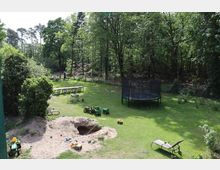 Grüne Gartenlandschaft mit Sandkasten, Spielzeugtraktoren, einer großen schwarzen Trampolinanlage und Sitzmöbeln auf einer Rasenfläche, umgeben von hohen Bäumen am Rand eines Waldes. Im Hintergrund stehen mehrere Tische mit Stühlen unter freiem Himmel.