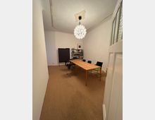 Ein einfacher Meetingraum mit einem langen rechteckigen Holztisch und mehreren schwarzen Stühlen, einem schwarzen Flipchart an der Wand und einem Regal mit Büchern und Ordnern. Eine große, kugelförmige Deckenlampe hängt zentral im Raum, der mit hellem Teppichboden ausgelegt ist.