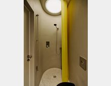 Kleine, moderne Dusche mit abgerundeten Wänden, einem runden Oberlicht und einem halbrunden, gesprenkelten Duschboden mit Ablauf. An der Wand sind eine Handbrause, eine eingelassene Ablagenische und ein gelber Duschvorhang zu sehen.