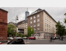 Straßenecke mit einem großen mehrstöckigen Wohn- oder Bürogebäude in heller Fassade und einigen begrünten Flächen, darunter ein Gebäudeteil mit Glasfenstern und Efeubewuchs. Im Hintergrund ragt der Turm einer Kirche oder eines Glockenstuhls empor, während im Vordergrund geparkte Autos, ein Fahrradfahrer und Passanten zu sehen sind.