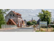 Straße in Bad Wilsnack mit historischen Backsteinhäusern und Bäumen am Straßenrand, im Hintergrund die Spitze einer Kirche. Im Vordergrund ein Schild mit der Aufschrift „Willkommen im Thermalsole- und Moorheilbad Bad Wilsnack“.