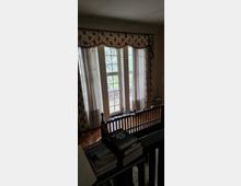 Ein Wohnraum mit großen bodentiefen Fenstern, die von gemusterten Vorhängen mit floralen Motiven eingerahmt sind. Im Vordergrund befindet sich ein hölzernes Geländer, und auf einer Ablage liegen Bücher; außen vor den Fenstern sind Bäume sichtbar.