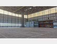 Große industrielle Halle mit hoher Decke und Metallkonstruktionen, durchsetzt von großen, rechteckigen Fenstern, die diffuses Tageslicht hereinlassen. Im Vordergrund ein leerer Betonboden; im Hintergrund befinden sich eine graue Büroeinheit und orangefarbene Industrietore.