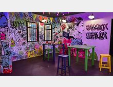 Ein Raum mit bunt gestalteten Wänden, bedeckt von Graffiti und Kunstwerken, darunter eine Frauenskizze und der Schriftzug „Paradox Berlin“. Im Vordergrund stehen bunte Tische und Hocker, während bunte Wimpelketten von der Decke hängen.
