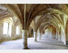 Gotischer Gewölberaum mit gemauerten Rundsäulen und spitz zulaufenden Gewölbebögen aus roten Ziegeln, umgeben von Steinwänden mit hohen Rundbogenfenstern. Der Boden ist gepflastert, und im Hintergrund führt eine Türöffnung in einen weiteren Raum.