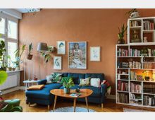 Wohnzimmer mit gemütlichem, dunkelblauem Sofa, dekoriert mit bunten Kissen, und einem kleinen Holztisch. An einer warm-orangenen Wand hängen gerahmte Bilder, daneben ein Regal mit Büchern, Zimmerpflanzen und Dekoration; links große Fenster, die viel Tageslicht hereinlassen.