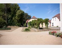 Ein ländlicher Hof mit hellen Gebäuden und roten Ziegeldächern, umgeben von Grünflächen und Bäumen. Im Vordergrund verläuft ein Kiesweg, daneben befinden sich bepflanzte Beete und eine mit Pflanzen bewachsene Pergola.