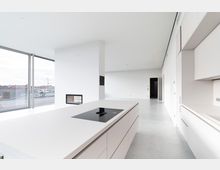 Minimalistisch gestalteter Innenraum einer modernen Küche mit weißen Schränken und einer zentralen Kücheninsel mit integriertem Kochfeld. Große Glasfenster bieten Ausblick auf Dächer und umliegende Gebäude, während eine eingebaute Wandöffnung zu einem angrenzenden Raum führt.