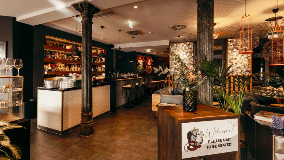 Innenraum einer stilvollen Bar oder eines Restaurants mit langem Tresen, Barhockern und beleuchteten Regalwänden voller Flaschen. Dunkle Säulen, warme Pendelleuchten und Pflanzen rahmen einen Empfangstisch mit „Welcome“-Schild im Vordergrund ein.