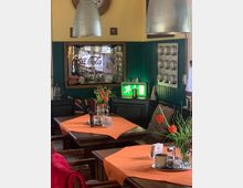 Ein gemütliches Restaurantinterieur mit Holztischen, die mit orangefarbenen Tischdecken und Vasen mit Blumendekoration dekoriert sind. Im Hintergrund hängen ein antiker Coca-Cola-Spiegel, Metalllampen sowie ein grün leuchtendes Notausgangsschild, umgeben von dekorativen Wandelementen.