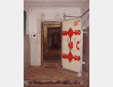 Betonierter Bunkerflur mit einer massiven Metalltür im Vordergrund, die mit roten Schließmechanismen ausgestattet ist und die Zahl „32“ trägt. Der Flur führt durch mehrere offene Türen in die Tiefe, während die Umgebung schlicht und funktional wirkt.