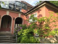 Roter Putzbau mit Rundbögen am Eingangsbereich, flankiert von blühenden Rhododendron-Büschen und Bäumen; darüber ein verglaster Balkon, im Hintergrund ein Ziegeldach und umliegendes Grün.