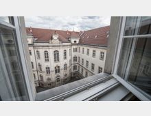 Blick aus einem geöffneten Fenster auf einen historischen Innenhof mit hellen Fassaden, zahlreichen Sprossenfenstern und einem roten Ziegeldach. Die Architektur zeigt klassische Elemente wie Bögen und verzierte Details.