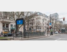 U-Bahn-Eingang mit der Beschilderung "Uhlandstraße" in einer städtischen Straßenlandschaft, umgeben von historischen Wohn- und Geschäftshäusern im klassischen Stil. Im Vordergrund eine Ampelkreuzung, Passanten und parkende Autos, im Hintergrund moderne und alte Architektur.