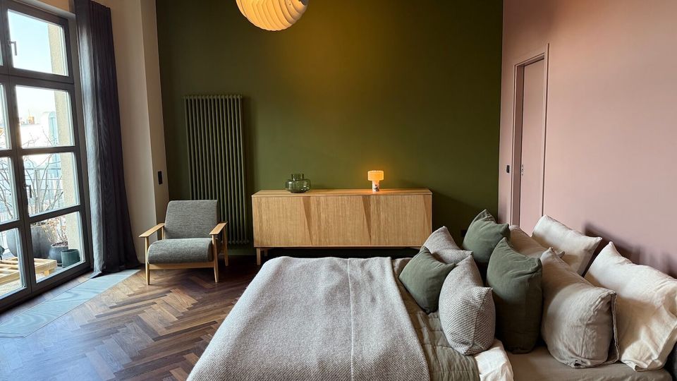 Modern bedroom with a large bed covered in neutral bedding and multiple pillows, set against a muted pink wall and a dark green accent wall. A wooden sideboard, tall radiator, gray armchair, and pendant light sit on herringbone wood flooring beside a large glass door to a small balcony.